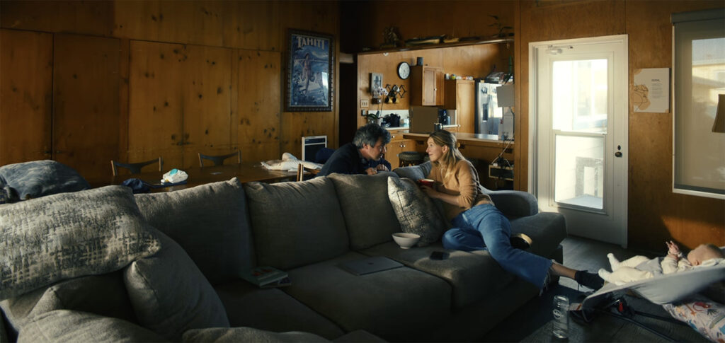 Nathan Fielder and Elizabeth Holmes sit on a couch in a cabin while she rocks a baby with her foot